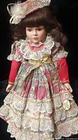 Porcelain doll in multi-layered pink floral and lace dress with matching bonnet and curly brown hair, showing detailed clothing and face.