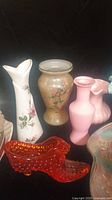 Photo showing a collection of vintage glass vases in various shapes and colors including a tall white vase with pink flowers, a frosted pink vase, and an ochre vase with floral painting.