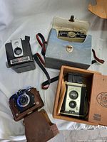 Photo showing four vintage cameras including Kodak Brownie in box, Polaroid Land Camera, a black box camera, and a blue leather camera.