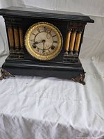 Front view of the Waterbury Clock Co. six-column mantel clock showing the black wooden case, six yellowish columns on each side, round brass bezel clock face with Roman numerals and minute markings, and decorative brass feet.