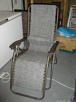 Folding antigravity lounge chair shown in sitting position, fabric seat and backrest attached to metal frame with brown armrests, photographed indoors on a concrete floor next to boxes and other household items.