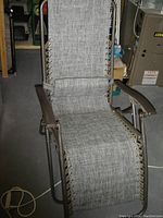 Folding zero gravity lounge chair with light gray woven fabric and metal frame, shown unfolded in a storage or workshop space with various items and equipment visible in the background.