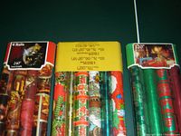 Photo showing three sealed packages of Christmas wrapping paper rolls with various festive patterns including ornaments, Santa Claus, candles, and holly.