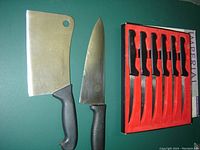 Photo shows 6 boxed steak knives with black handles in red holder, one large chef knife with black handle, and one Henckels cleaver with black handle and hole in blade