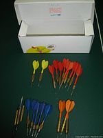 Photo of 18 darts grouped by flight color: 2 yellow, 10 red, 6 blue, and 4 orange darts laid out above a white open box.