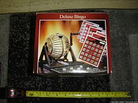 Photo of the Deluxe Bingo game box with bingo cage, balls, and cards displayed on top and a measuring tape below.