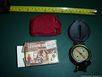 Top view of engineer's directional compass open to show compass face, with degree markings, magnetic needle, and sighting slot on lid. Includes measuring tape and red pouch next to compass. Original product box is partially visible, showing outdoor scenes and usage suggestions.