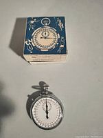 Mechanical stopwatch placed on flat surface next to its original blue and white illustrated box, showing the stopwatch face and box top with sports-themed graphics.