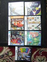 Seven Nintendo DS game cases displayed in a grid on a dark surface with a carpet visible. Titles include Polarium, Prism Light the Way, Art Academy, TouchMaster 2, Big Bang Mini, Mister Slime, and 101 in 1 Megamix. All cases have original cover art and are in protective plastic casing.