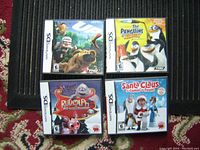 Photo showing four Nintendo DS game cases arranged side by side on a carpet. Titles visible: UP, The Penguins of Madagascar, Rudolph Red Nose Reindeer, and Santa Claus is Coming to Town.
