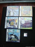 Top-down photo showing six Nintendo DS game cases arranged in two columns and a loose game cartridge to the side. Cases have artwork for Seaworld Shama's Deep Sea Adventures, Silly Bandz, High School Musical Making the Cut, Tomagotchi Connection Corner Shop 2, Defending the Penguin, and Winx games.