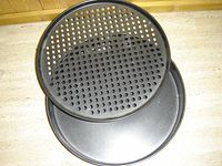Two black pizza pans, one solid and one with multiple circular perforations, both 14 inches in diameter, stacked one on top of the other on a wooden surface.