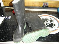 A pair of black rubber boots placed on a surface with one boot standing upright and the other lying horizontally showing the green treaded sole.