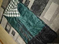 Photo of an Ozark sleeping bag spread out on floor, displaying green and black exterior with plaid interior lining.