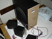 Image showing one large subwoofer next to four small black satellite speakers on a table with cables connected.