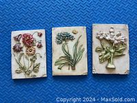 Three ceramic tiles each displaying detailed floral relief designs in pastel colors on cream backgrounds, arranged side by side on a blue textured surface.