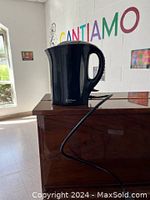 Black electric kettle resting on a wooden surface in a well-lit room with windows and colorful wall text.