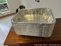 Stack of approximately 15 square disposable metal trays placed on a wooden surface, with light coming from a nearby window.