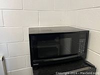 Front and side view of a black Danby microwave oven sitting on a black mini-fridge against a white brick wall.