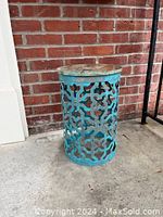 Blue metal plant stand with geometric cut-out pattern shown standing on a concrete floor near a red brick wall.