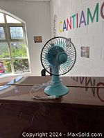 Teal USB desk fan shown from the front placed on a dark wood surface, with USB cable visible.