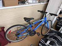 Side view of blue Minnelli Pro 24 18 inch bicycle with lock wrapped around frame. Bicycle positioned against wall showing tires, pedals, saddle, and kickstand.