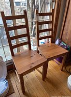 Two wooden high back dining chairs with ladder-style horizontal slats on the backrest, natural wood finish, visible wood grain and knots, sitting on wood flooring near a window.