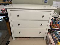 Front view of white dresser showing three drawers with black metal knobs and some visible marks on the finish.