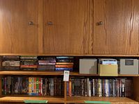 Three shelves of tightly packed DVDs, DVD box sets and CD cases on wooden shelves under cabinetry. DVDs include various titles and are mixed orientation. CDs stored in boxes on right.