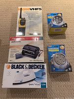 Full lot arranged on floor showing all boxed items: Humminbird VHF5 radio, SentrySafe AS-1 safe, Black & Decker iron, and two LED motion sensor lights.