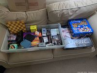 Photo of the assortment of games including chessboard, card games boxes, and game boards in an open plastic bin plus the boxed Scrabble and Disney Chess Set on couch.