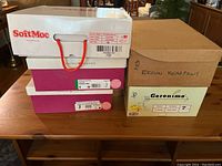Four shoe boxes stacked on a wooden table, showing brand labels for Soft Moc, Naturalizer, Geronimo, and Bearpaw