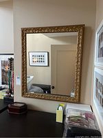 Front-facing photo of rectangular mirror hanging on beige wall, showing detailed gold ornate composite frame and reflections.
