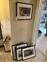 Photo showing five black and white photo prints, framed and matted, three leaning against wall, one hanging above stairwell.