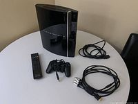 Shows PlayStation 3 console, remote control, wired controller, and cables on white round table.