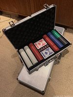 Open aluminum case displaying poker chip set with white, red, green, blue, black chips and two decks of playing cards, dice
