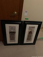 Two vertically oriented black frames leaning against a wooden door, each containing a black and white photo print of architectural details