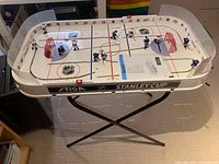 Photo of Stiga rod hockey table on black metal stand showing player rods, red goal nets, NHL logos on rink surface, and some visible damage near one goalie net.