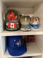 Photo of display shelf with three snow globes, including Canada-themed snow globe with flags and maple leaf, London Big Ben snow globe, and skiing winter scene snow globe, with blue Toronto Blue Jays baseball cap below