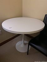 Round white table with metal pedestal base and laminated laminated white top, next to a black chair, showing full top and part of base