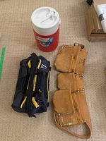 Photo showing red and white Coleman beverage cooler, navy and yellow Mastercraft tool bag, and tan Kunys leather tool belt laid out on carpet