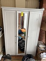 Front view showing three-door white wardrobe with central mirror door and black knob handles, surrounded by miscellaneous items.
