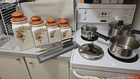 Photo showing three stainless steel pots with lids and four ceramic floral storage canisters with wooden lids sitting on kitchen countertop next to a white wire rack