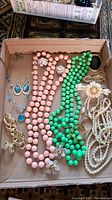 Photo of assorted costume jewelry in a box showing pink and green large bead necklaces, pearl-like strands, multiple bracelets and several pairs of earrings.