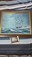 Framed oil painting of a tall ship with multiple sails on a wave-filled sea placed on a striped fabric background.
