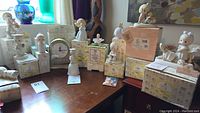 Overview of multiple boxed and unboxed Precious Moments porcelain figurines arranged on a table, showing a variety of designs including a clock, girls holding animals and other sentimental children figures.
