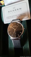 Close-up of Skagen wristwatch with black dial and silver markers, visible warranty card in background.