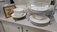 View of white dishes arranged on a countertop, including stacked plates, bowls, two metal trivets, a metal wire shelf with dishes underneath, and 4 clear glasses upside down.
