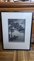 Framed black and white watercolour painting showing a person sitting in a boat with trees and buildings in the background.