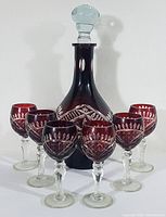 Front view of deep ruby red cut to clear Bohemian crystal decanter with six matching cordial glasses arranged in a semicircle around it, clear faceted stopper on decanter visible.